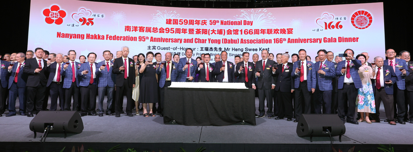 Group of people in suits raising wine glasses on stage, with large banner for Nanyang Hakka Federation event.